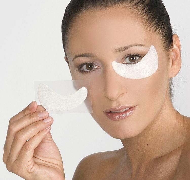 Nahaufnahme einer Frau mit Augenpads. Ein Pad wird gehalten, eines ist unter dem Auge. Weiße Pads.