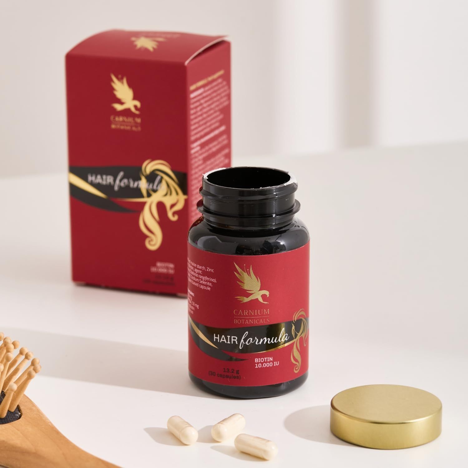 Flasche und Karton mit rotem Etikett. Text: CARNIUM BOTANICALS Hair Formula. Goldener Deckel. Kapseln liegen daneben.