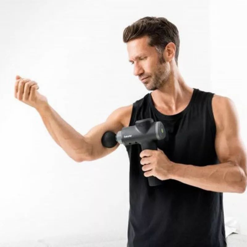 Mann verwendet Massagegerät am Arm. Schwarzes Massagegerät mit grauem Aufsatz. Mann trägt schwarzes Tanktop. Weißer Hintergrund.