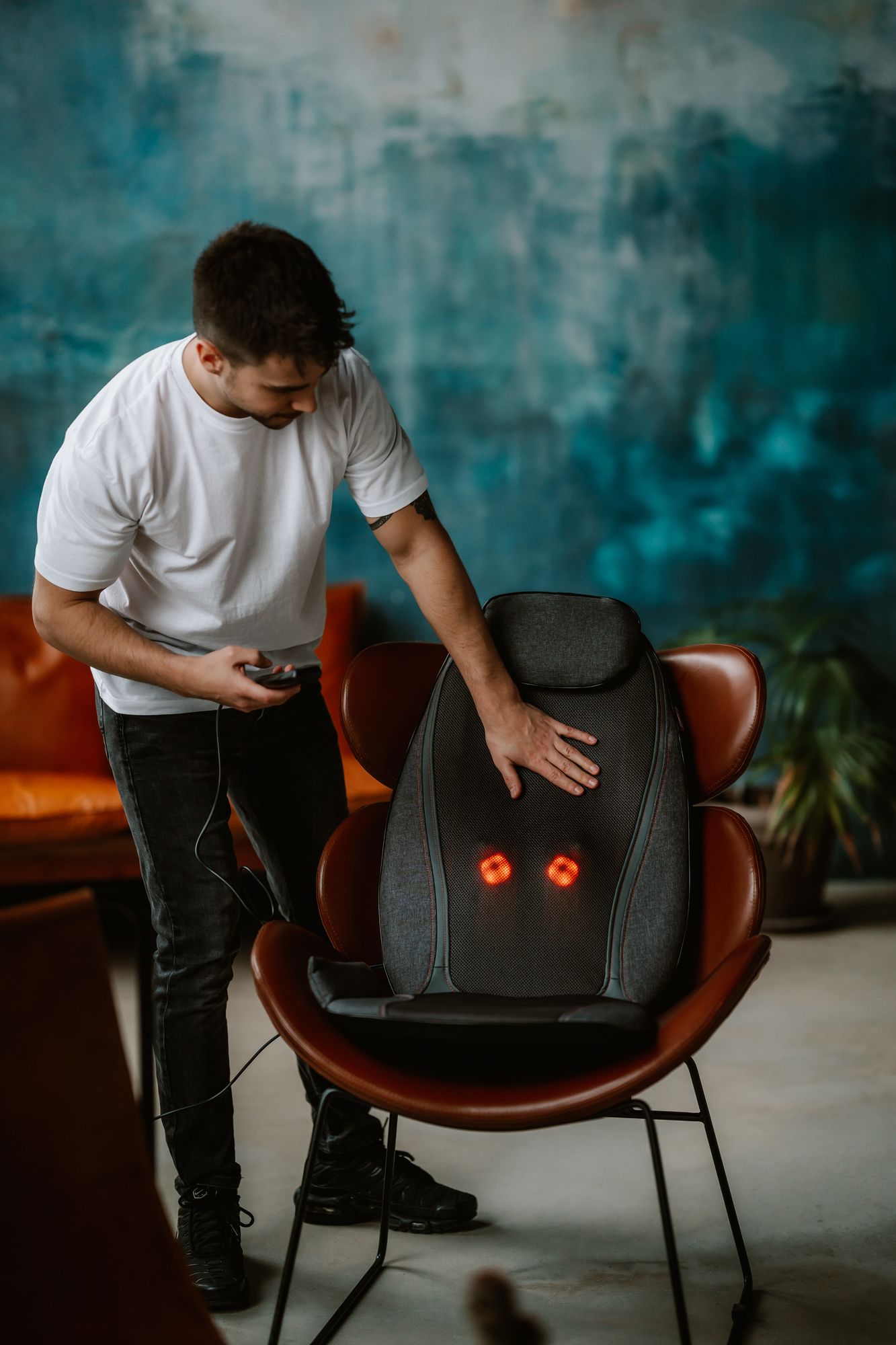 Mann mit weißem T-Shirt und Jeans berührt eine Shiatsu-Massagematte auf einem Stuhl. Rote Lichter sind sichtbar.