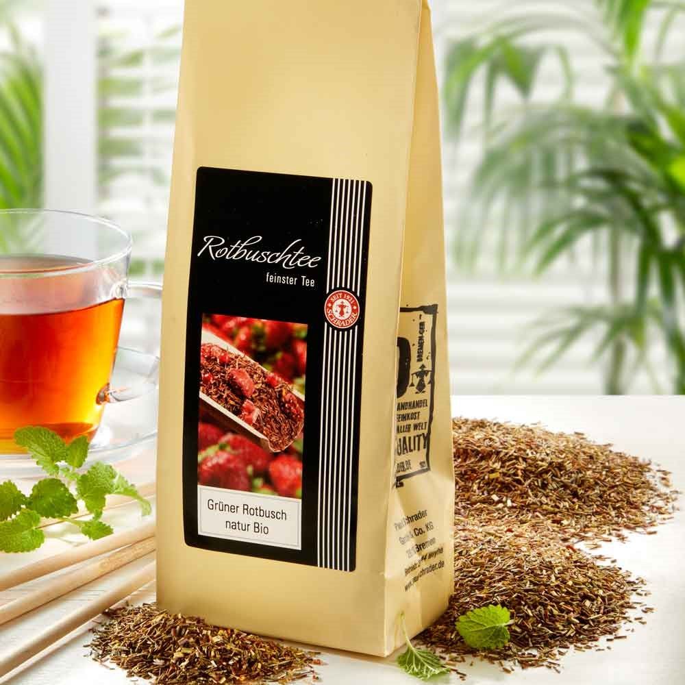 Schrader Grüner Rooibostee Natur Bio in Verpackung. Tee daneben verstreut. Glas Tee im Hintergrund. Bio-Siegel.