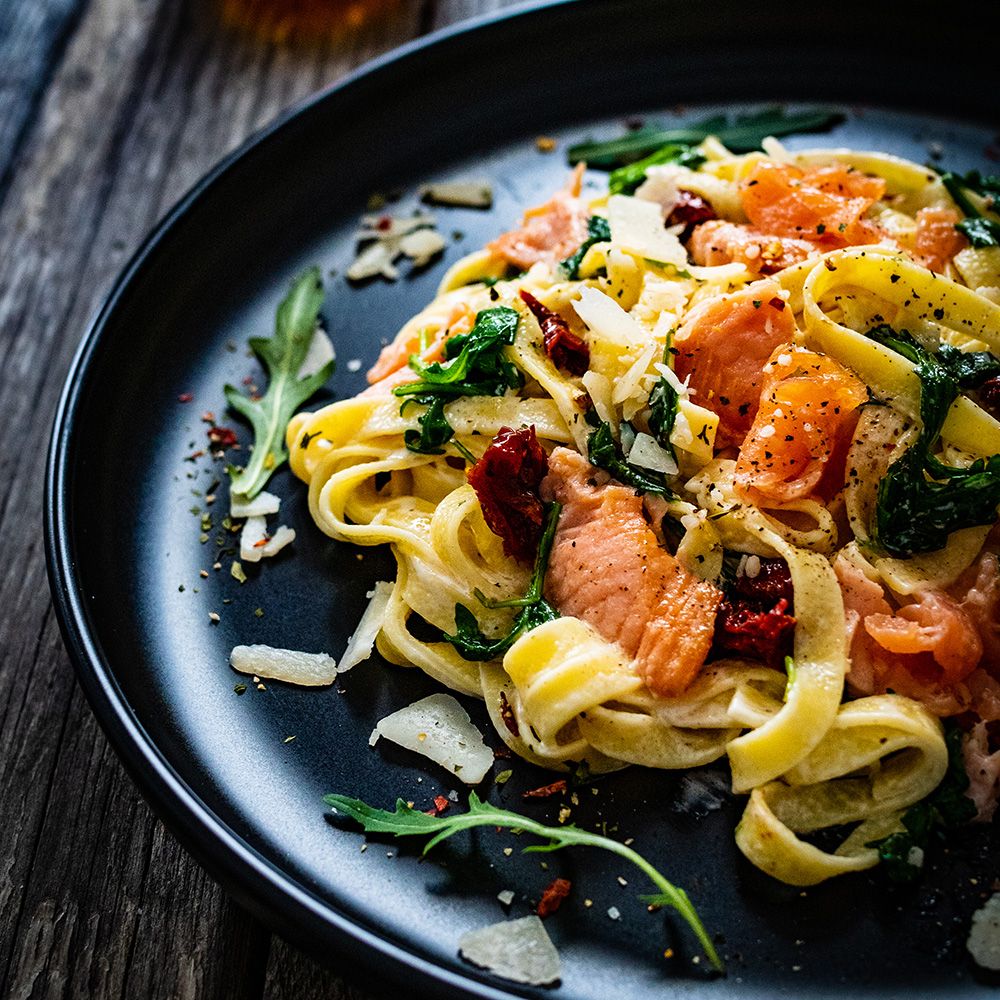Pasta mit Lachs, getrockneten Tomaten, Rucola und Parmesan auf einem schwarzen Teller. Holzuntergrund.