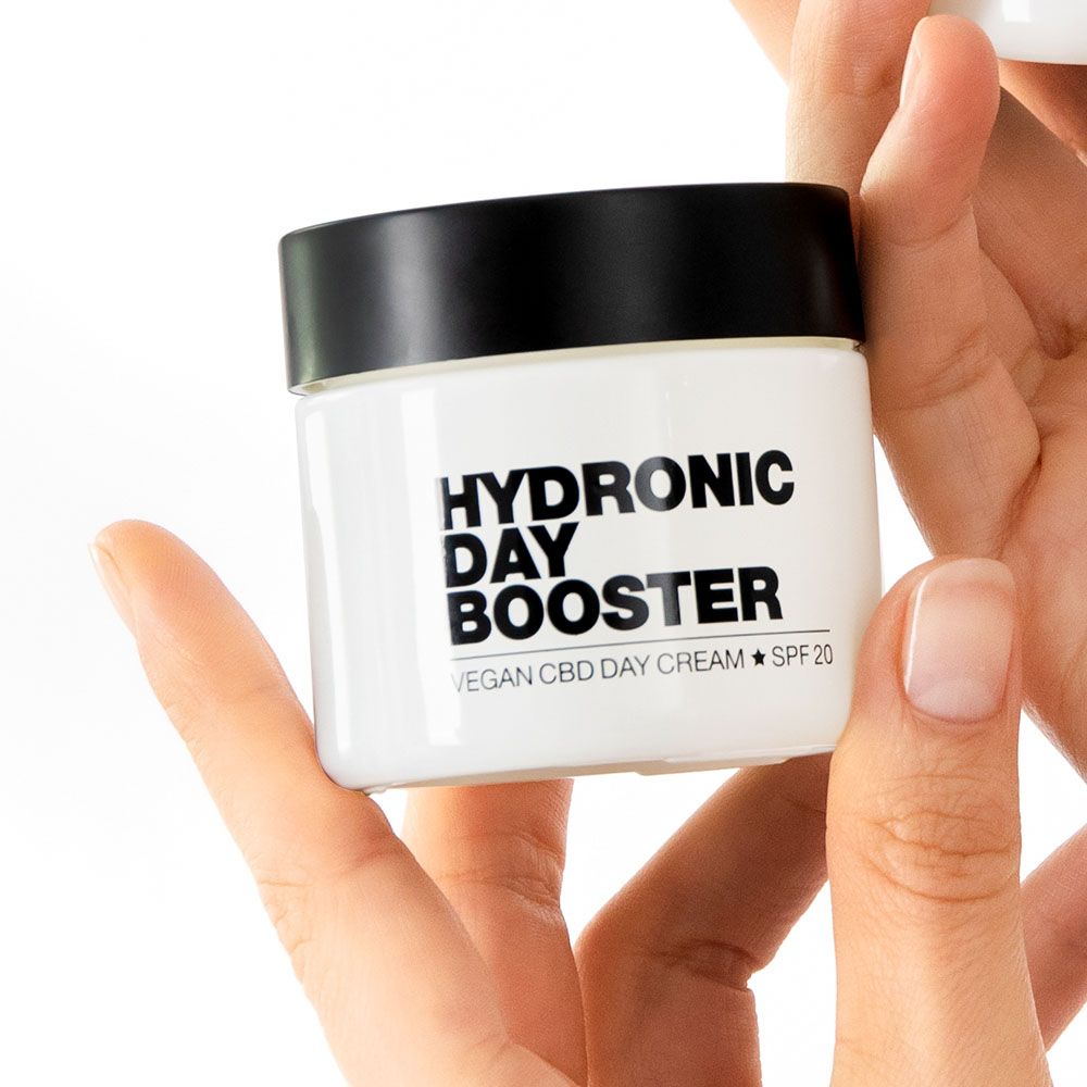 Weißer Tiegel mit schwarzem Deckel, gehalten von Händen. Aufschrift: Hydronic Day Booster, Vegan CBD Day Cream, SPF 20.