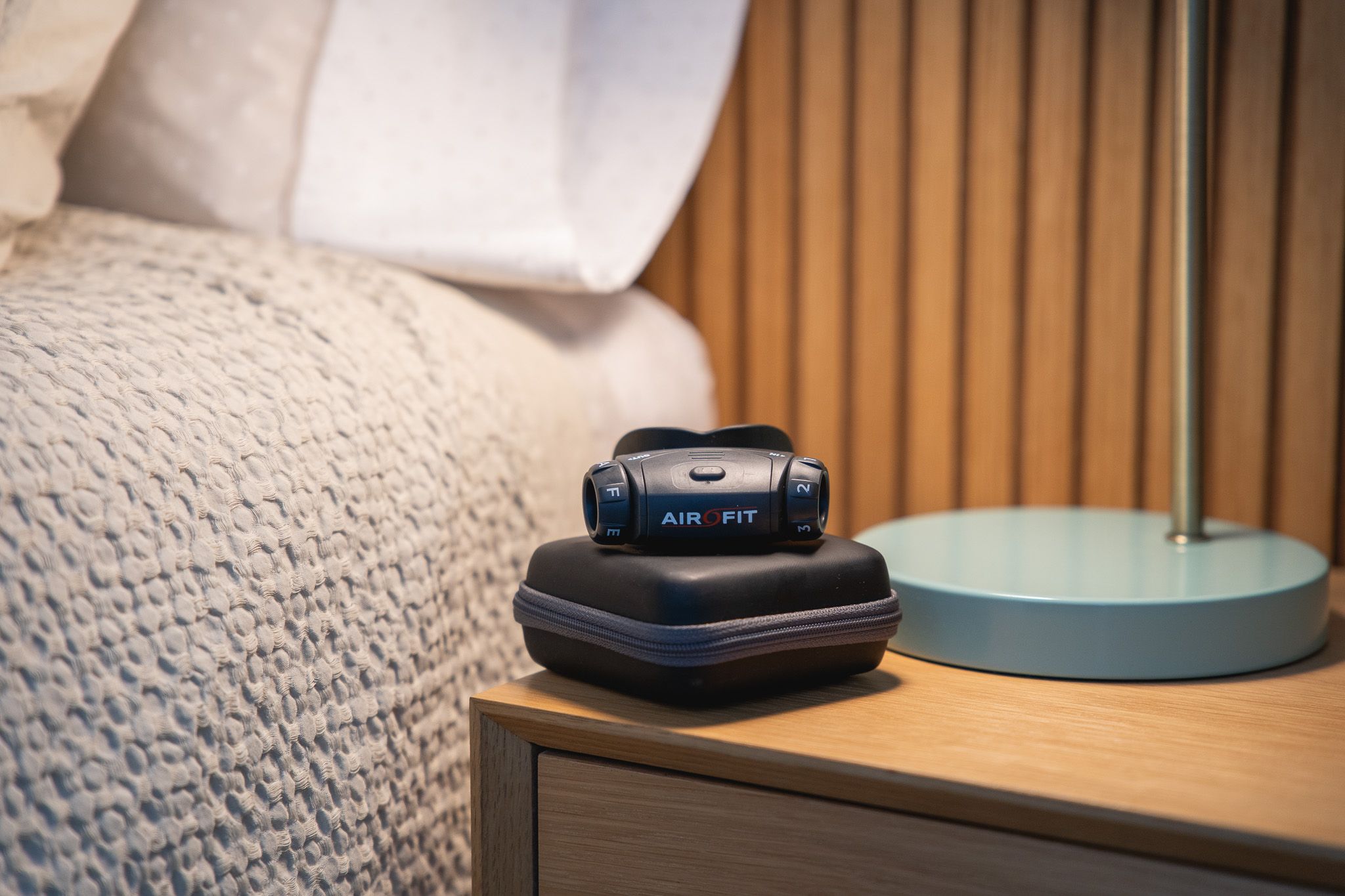 Schwarzes Atemtrainingsgerät auf einer schwarzen Box. Auf einem Nachttisch. Im Hintergrund Bett und Lampe.