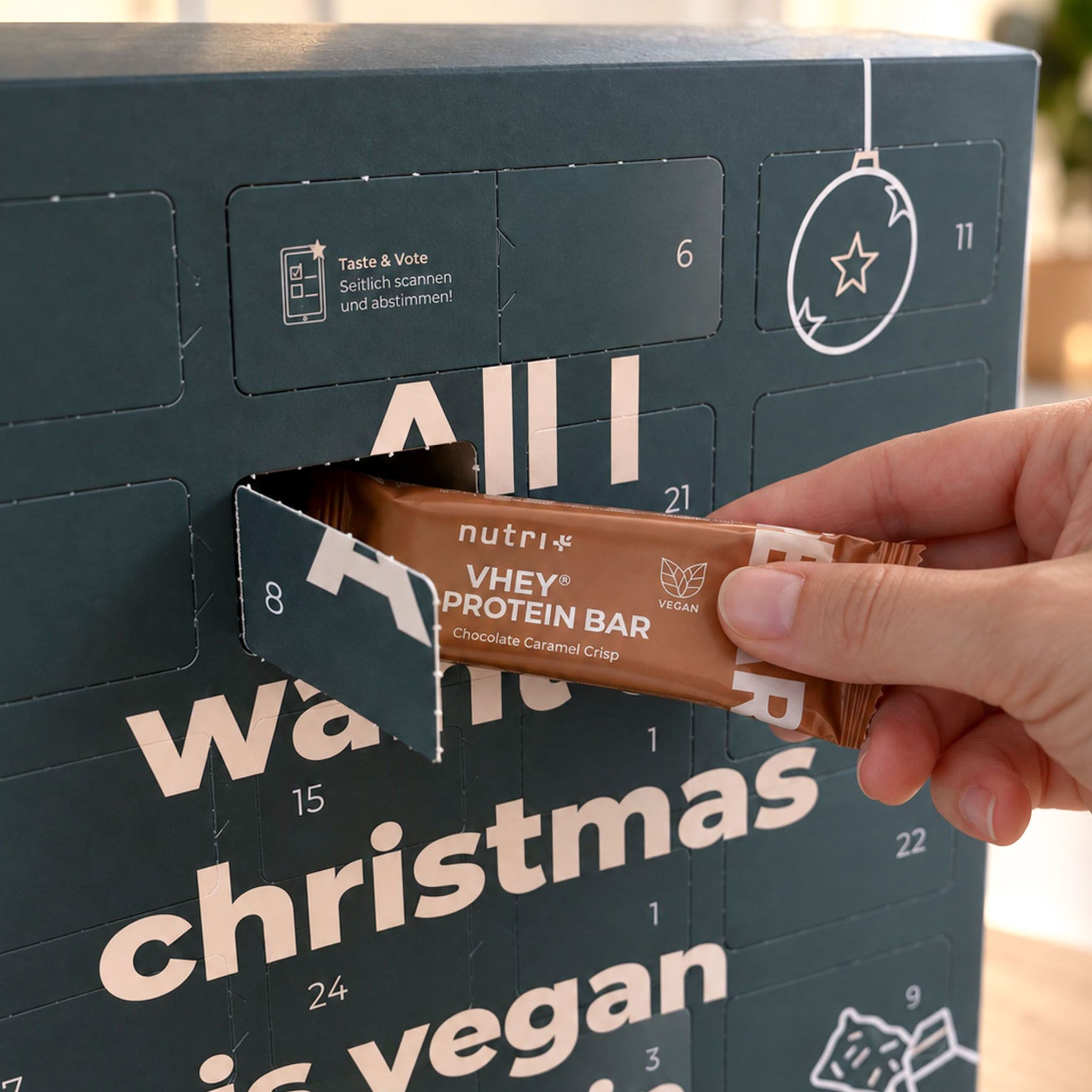 Hand öffnet eine Tür eines Adventskalenders und entnimmt einen Nutri-Vhey Proteinriegel, Schoko Karamell Crisp.
