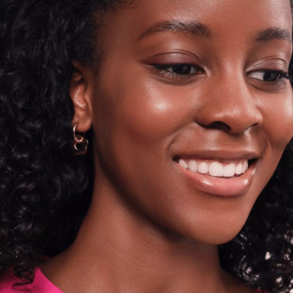 Porträt einer lächelnden Frau mit Locken und goldenen Ohrringen. Sie trägt rosa Lippenstift.