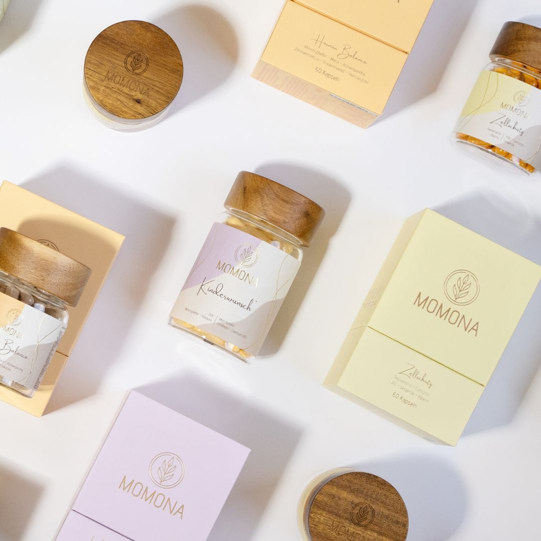 Mehrere Momona-Produkte: Gläser und Schachteln. Aufschrift Momona und Zellschutz. Holzdeckel und gelbe, rosa Schachteln.