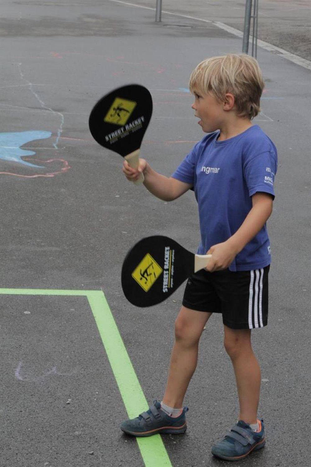 Kind hält zwei Street Racket Schläger. Schwarze Schläger mit gelbem Logo. Kind trägt blaues T-Shirt und schwarze Shorts.