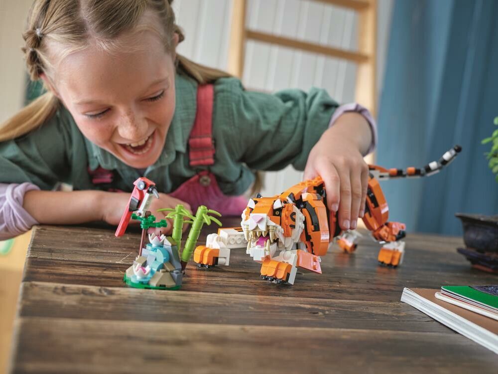 Un bambino gioca con un set LEGO Creator 3 in 1 che rappresenta una tigre. La tigre è arancione e nera. Accanto alla tigre si vedono un uccello rosso e una costruzione più piccola. Il bambino sorride.