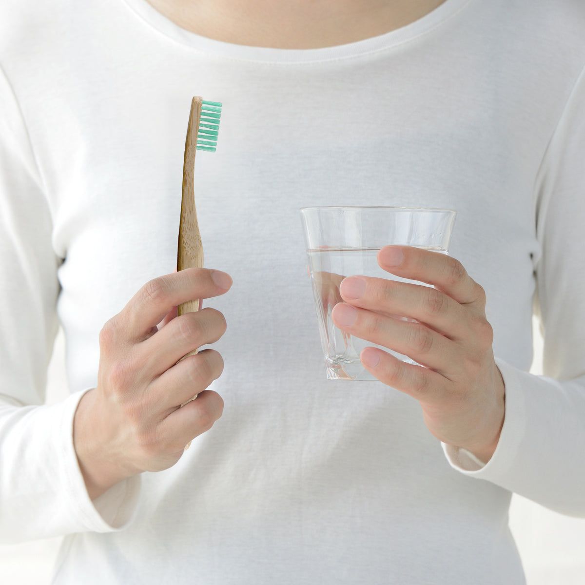 Person hält Bambus-Zahnbürste mit grünen Borsten und ein Glas Wasser. Person trägt weißes Oberteil.