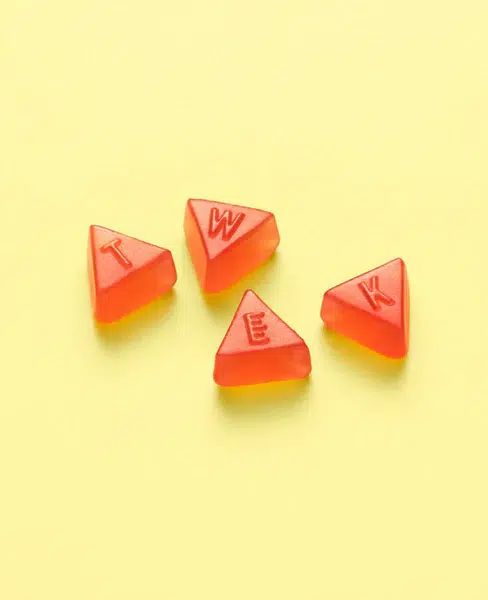 Vier dreieckige, orangefarbene Fruchtgummis mit Buchstaben. Auf jedem Gummi ist ein Buchstabe des Wortes TWEK eingeprägt.