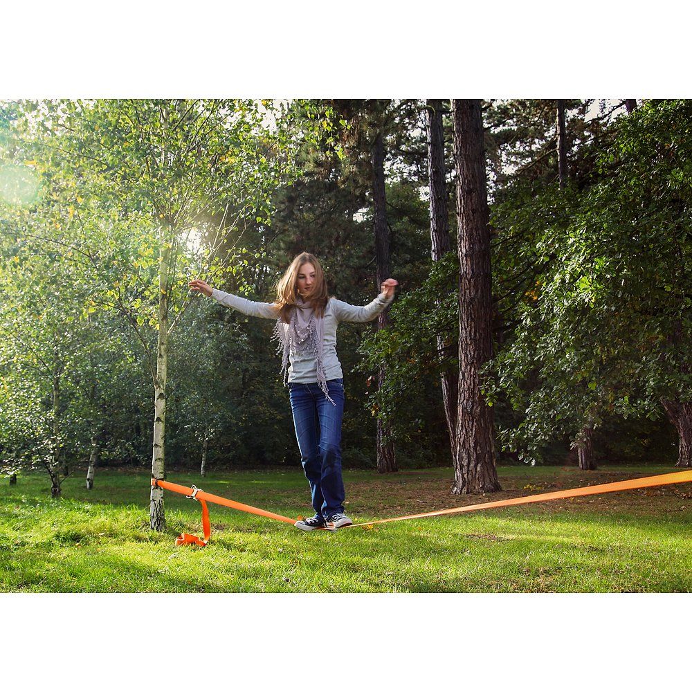 Frau balanciert auf einer orangefarbenen Slackline im Freien, befestigt zwischen zwei Bäumen.
