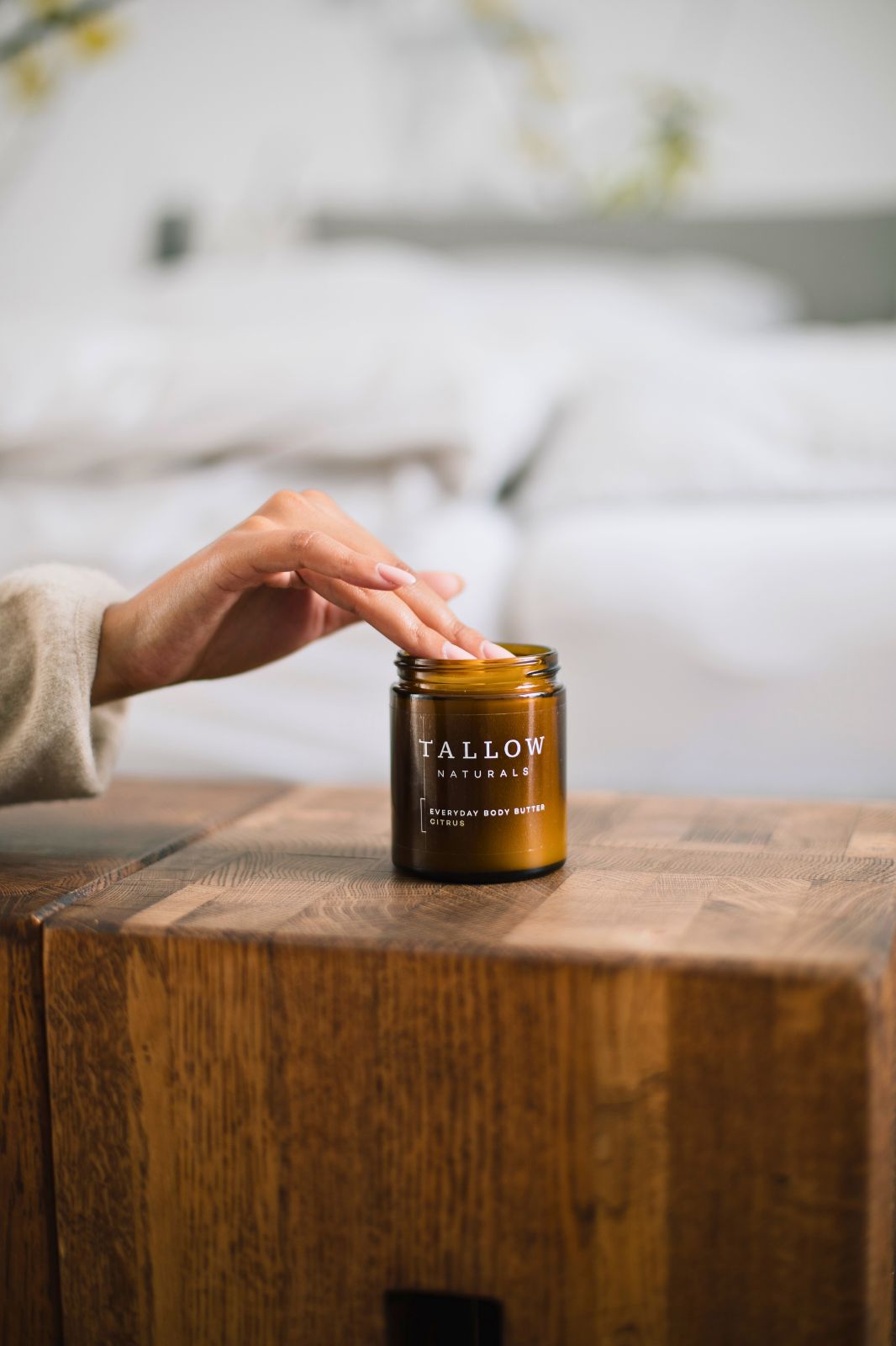 Hand berührt eine braune Dose mit schwarzem Deckel. Aufschrift: TALLOW NATURALS, EVERYDAY BODY BUTTER CITRUS. Dose steht auf Holz.