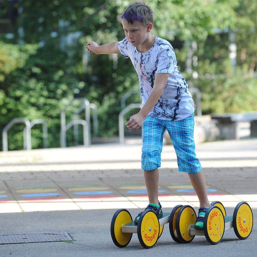 Kind auf Pedalo Koordinationstrainer. Gelbe Räder, Holzrahmen. Aufschrift: Pedalo. Das Kind bewegt sich.
