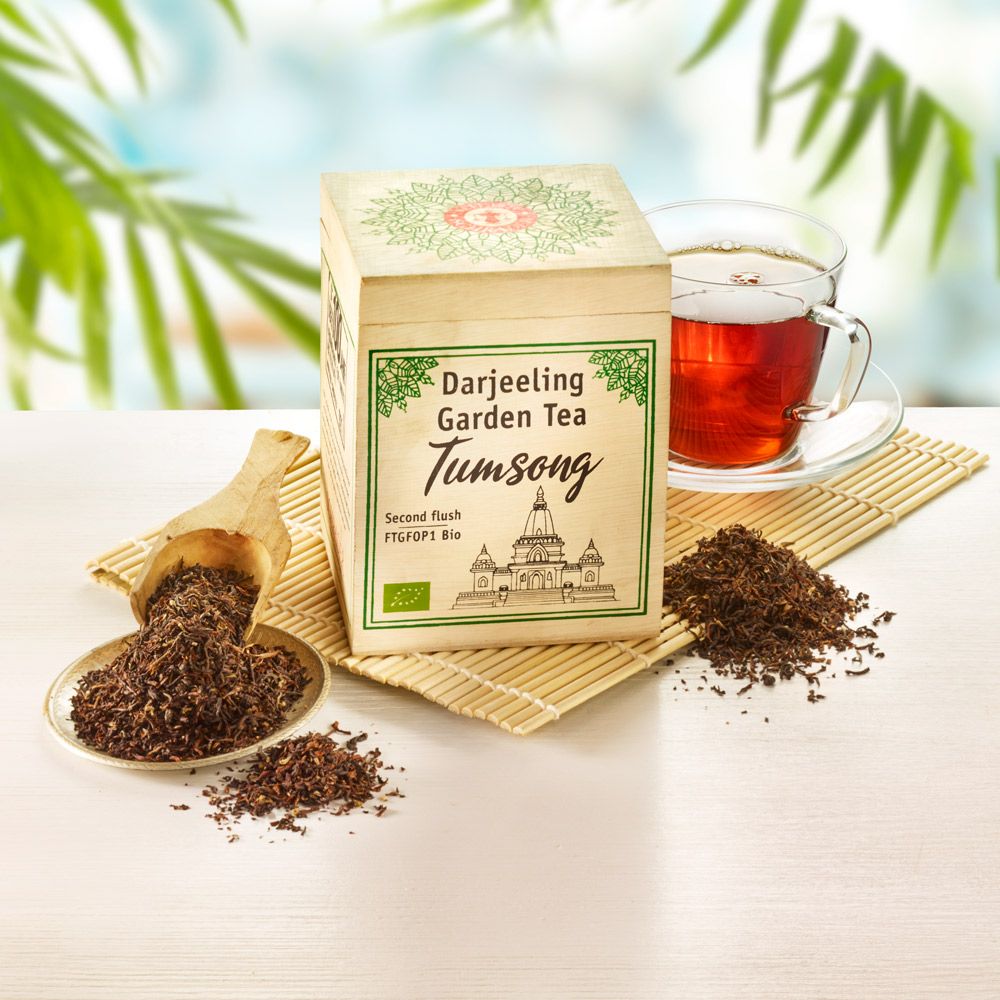 Holzkiste mit 'Darjeeling Garden Tea Tumsong'. Tee, Holzlöffel, Tasse Tee. Bio-Siegel. Auf Bambusmatte, heller Hintergrund.