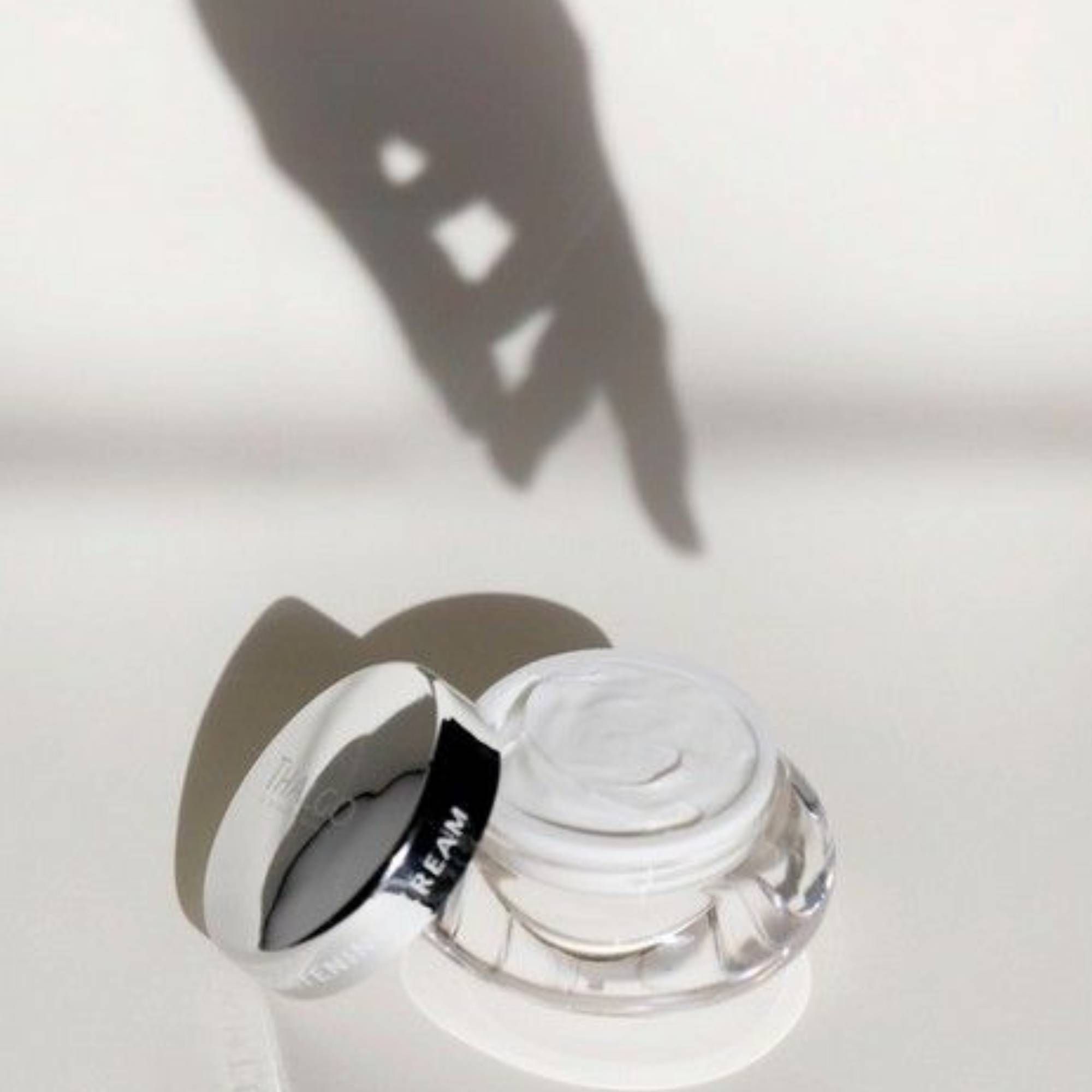 Offener Tiegel mit silbernem Deckel. Aufschrift: CREAM. Schatten einer Hand im Hintergrund.