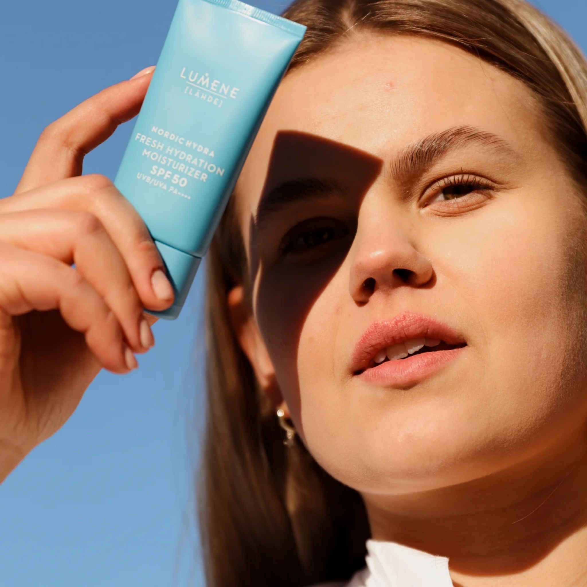 Frau hält hellblaue Tube vor blauem Himmel. Aufschrift: LUMENE [LÄHDE], Nordic Hydra Fresh Hydration Moisturizer SPF50. Gesicht im Schatten.
