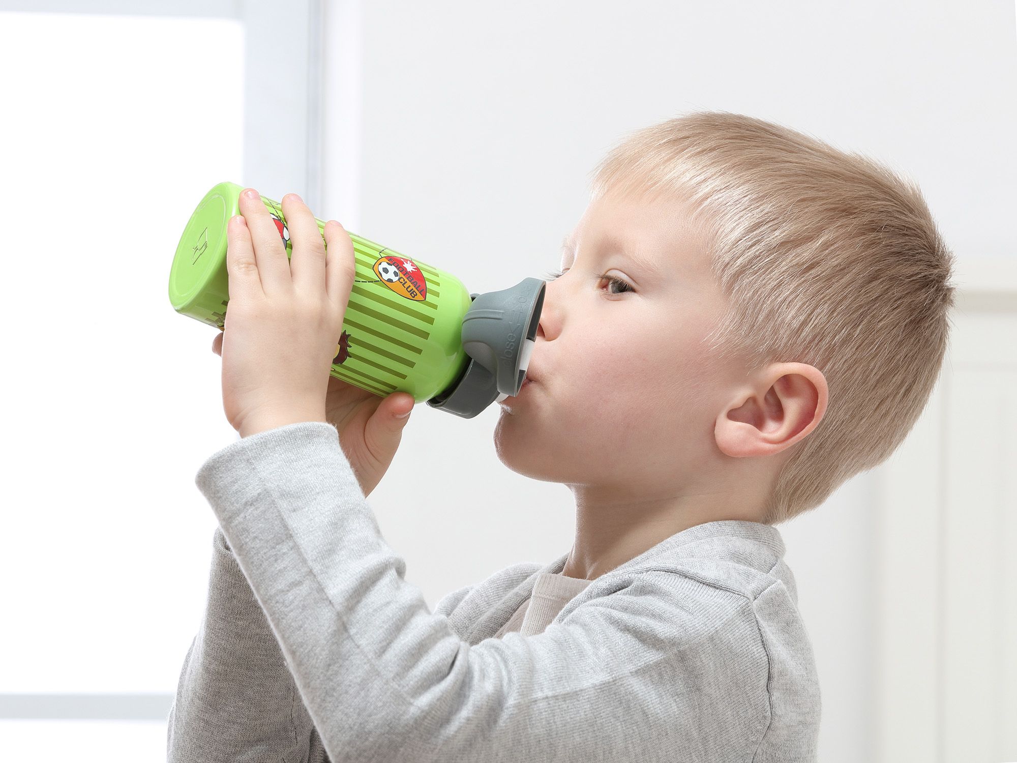 Kind trinkt aus grüner Trinkflasche. Flasche mit Fußball- und Streifenmuster. Kind hält Flasche mit beiden Händen.