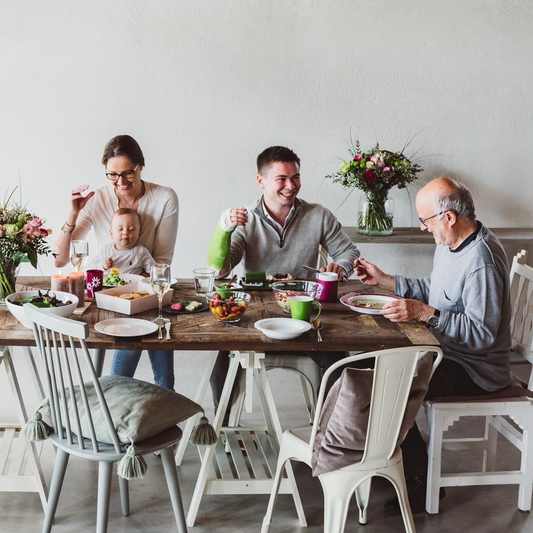 Familie sitzt an einem Tisch. Personen halten Becher. Auf dem Tisch Essen und Blumen. Ein Mann hat einen grünen Verband.