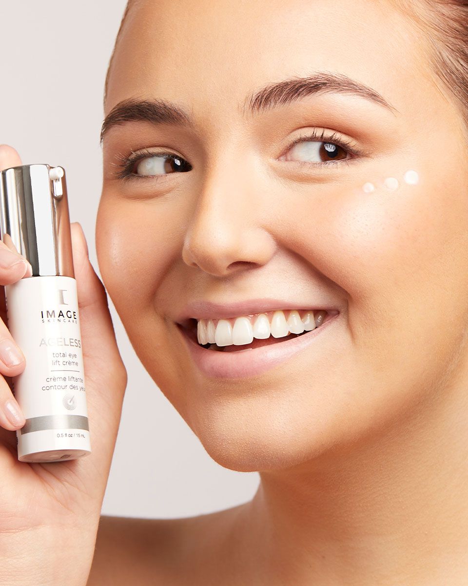 Frau hält eine Flasche. Aufschrift: IMAGE Skincare, AGELESS total eye lift crème. Creme-Punkte unter dem Auge.