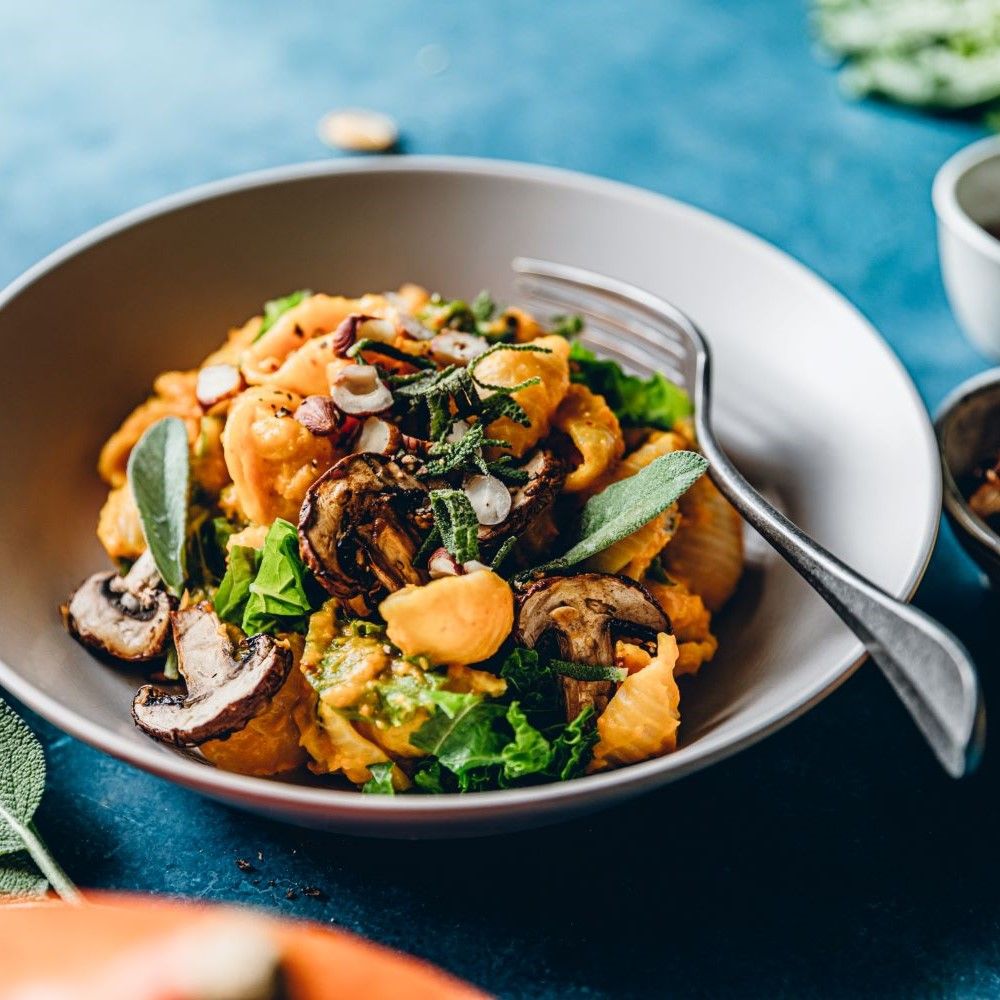 Schüssel mit Pasta, Kürbis, Pilzen und Salbei. Auf blauem Untergrund. Gabel liegt daneben. Dekoration.