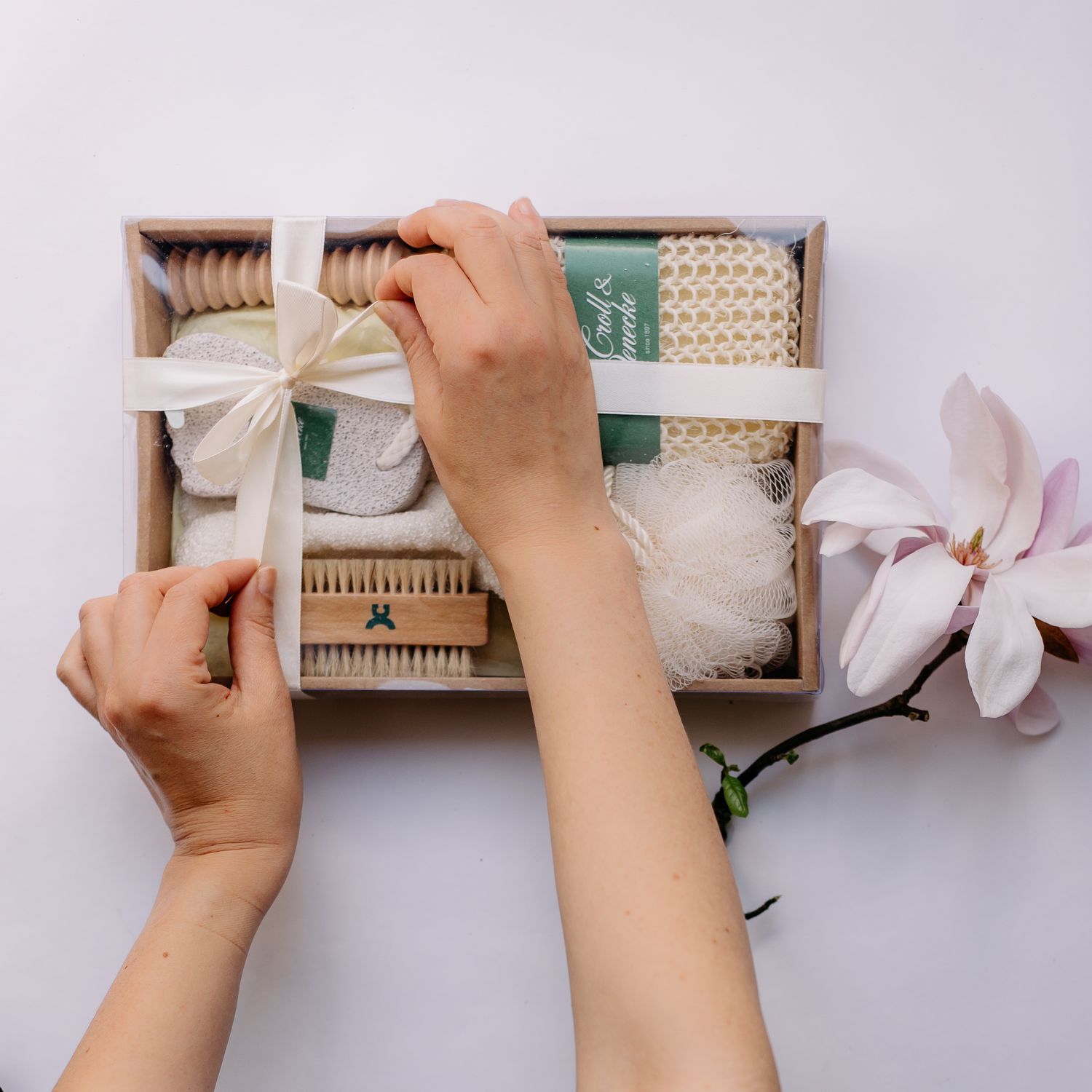 Geschenkset SPA: Hände binden eine weiße Schleife um eine Holzbox mit Pflegeprodukten wie Bürste und Schwamm.