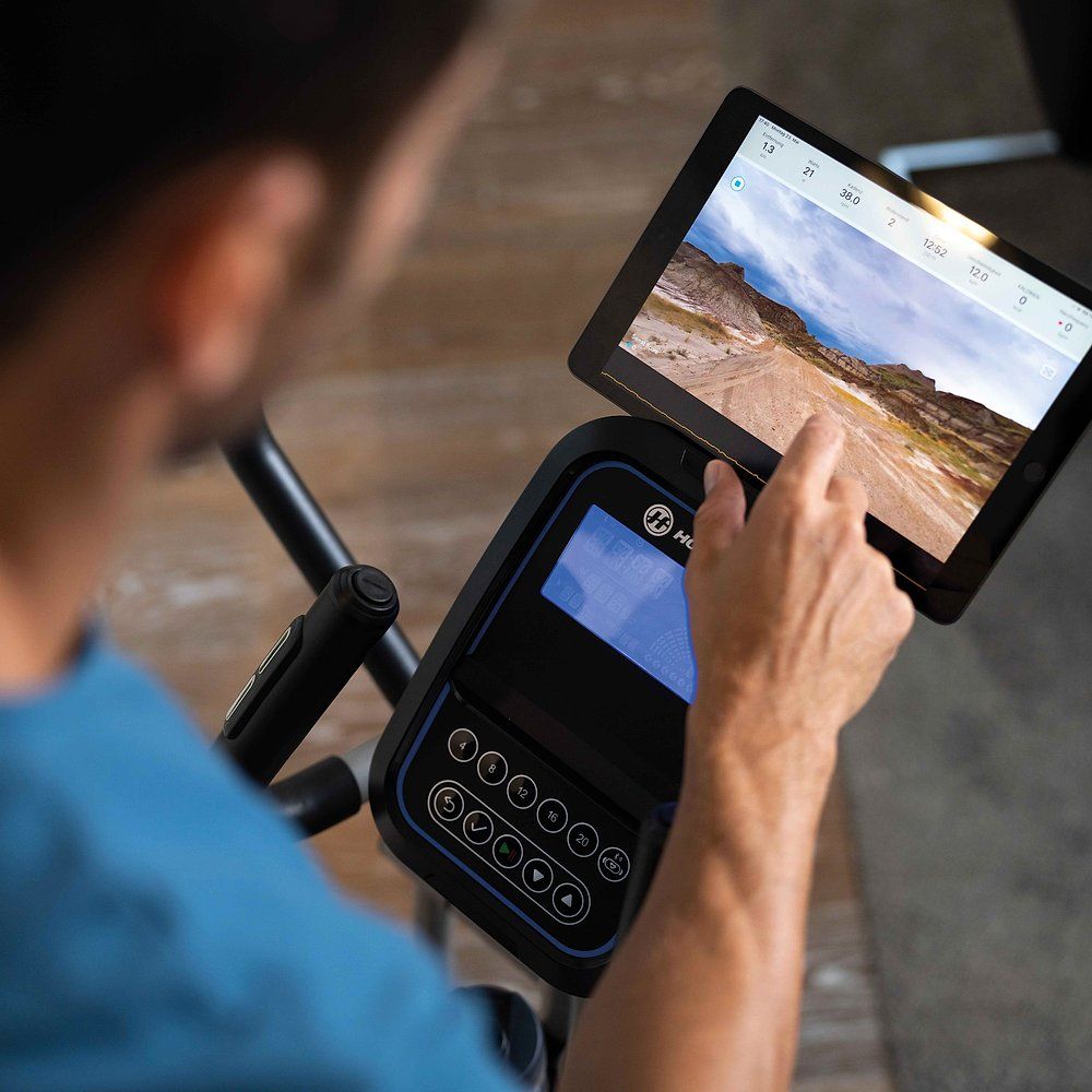 Person bedient Tablet auf Crosstrainer. Tablet zeigt Landschaft. Schwarzes Bedienfeld mit Knöpfen. Im Hintergrund ein Raum.