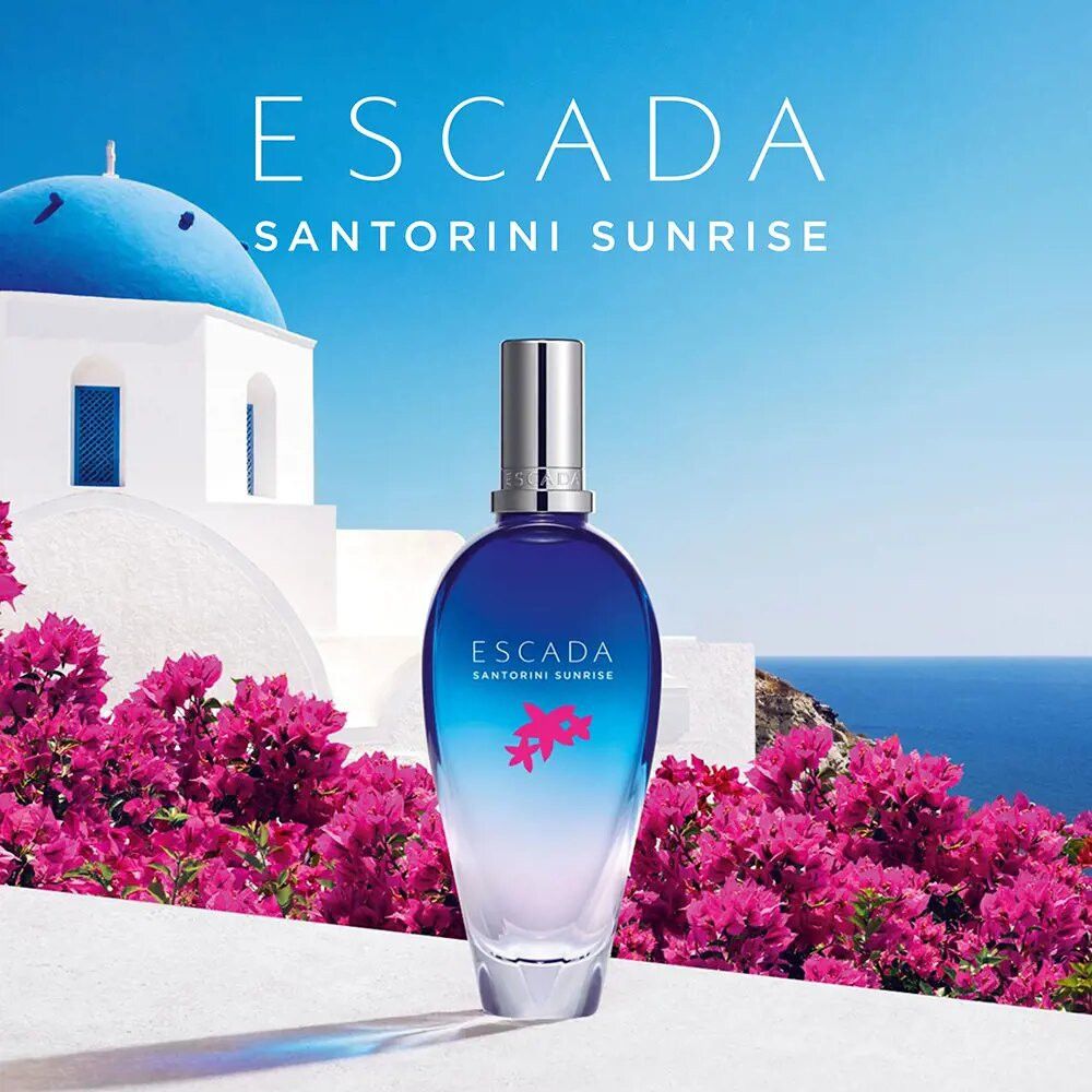 Escada Santorini Sunrise Flakon vor blauem Meer und Blumen. Blauer Flakon, silberner Deckel. Hintergrund mit Blumen und Meer.