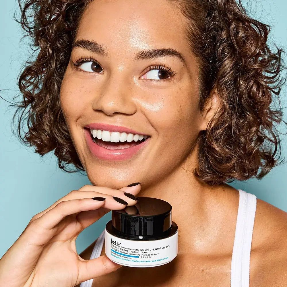 Lächelnde Frau mit lockigem Haar hält einen Tiegel Belif The True Cream Aqua Bomb.