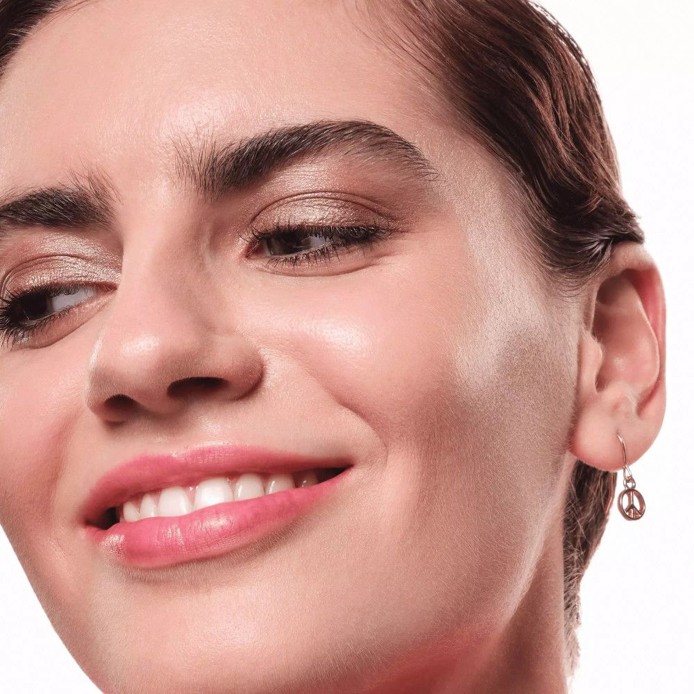 Nahaufnahme einer Frau mit braunem Lidschatten. Sie lächelt. Ein Ohrring mit Friedenszeichen.