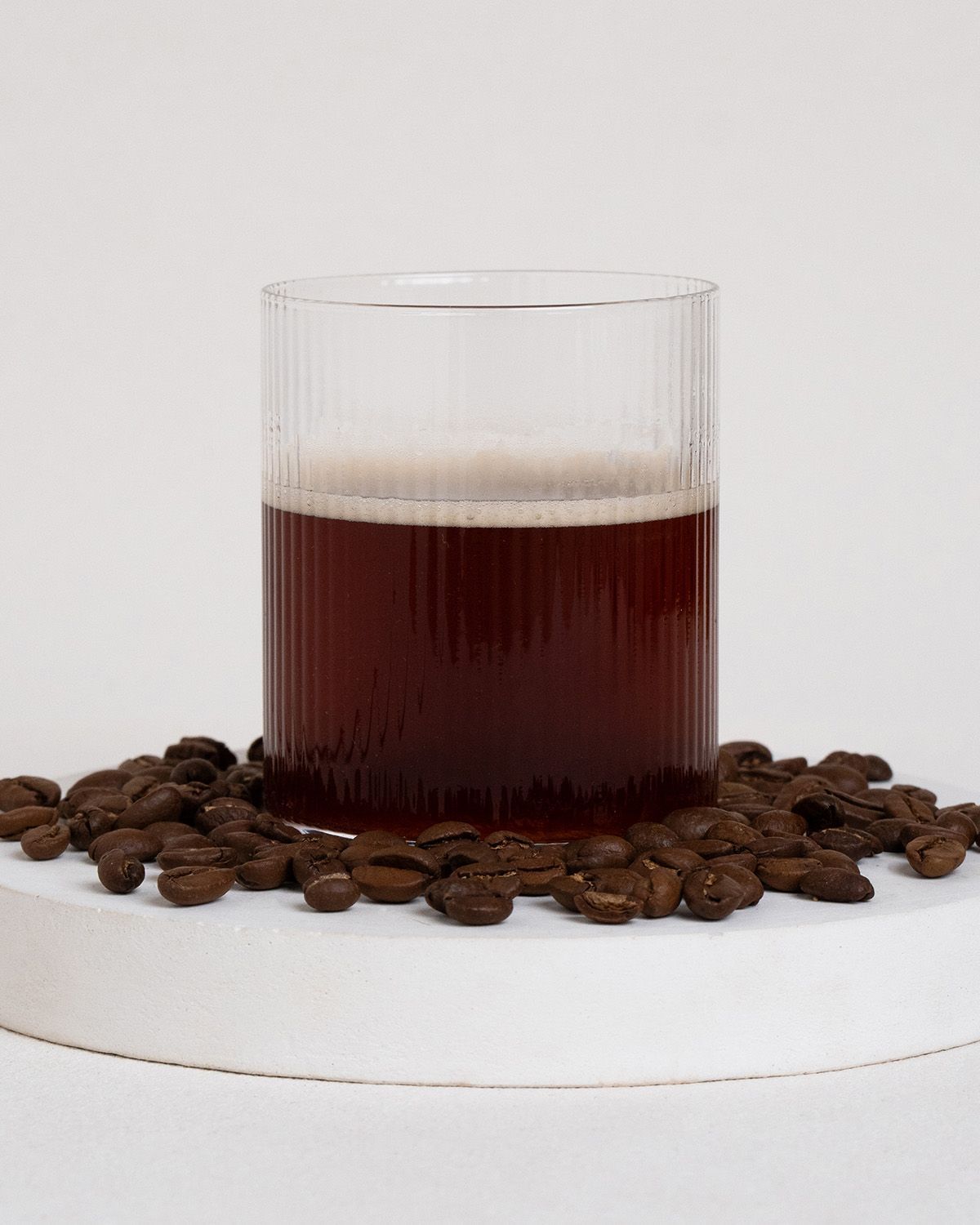Glas mit dunklem Kaffee und Milchschaum, umgeben von Kaffeebohnen.