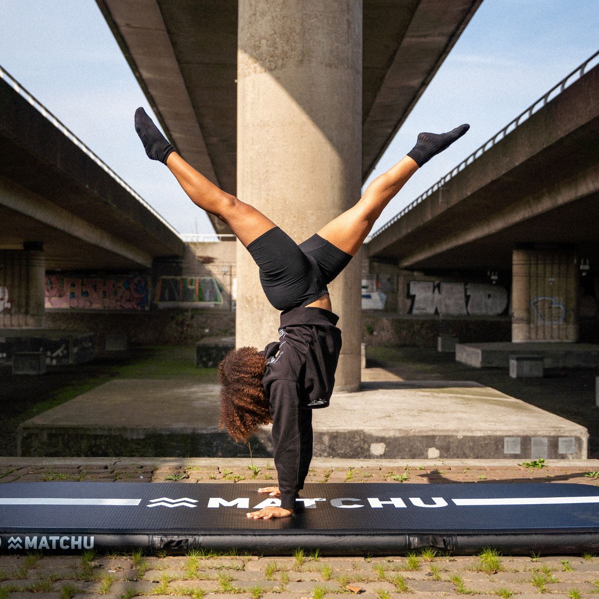 Person macht Handstand auf schwarzer Gymnastikmatte. Marke: MATCHU. Hintergrund: Betonkonstruktion.