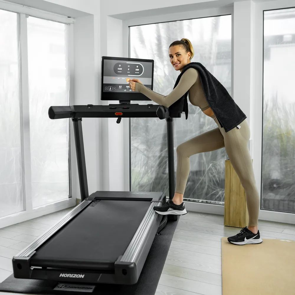 Femme debout à côté d'un tapis de course. Elle s'appuie sur le tapis de course. Tapis de course noir. Logo Horizon visible.