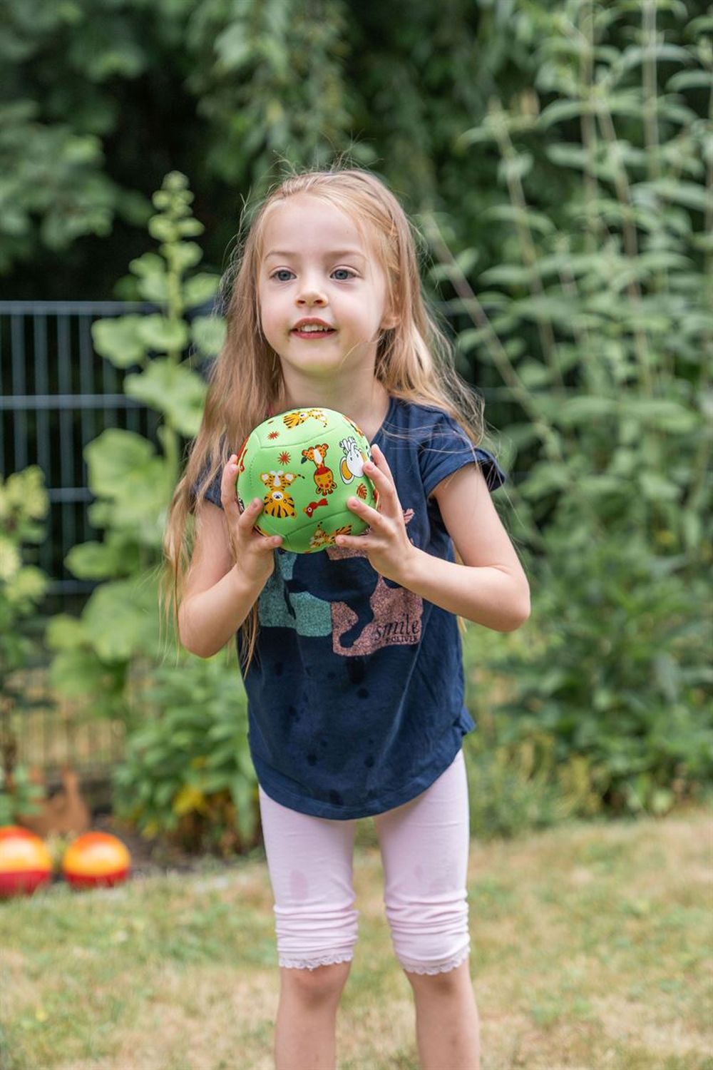Ein Mädchen hält einen grünen Softball mit Tier-Illustrationen. Das Mädchen steht in einem Garten. Sie trägt ein blaues T-Shirt und rosa Leggings.