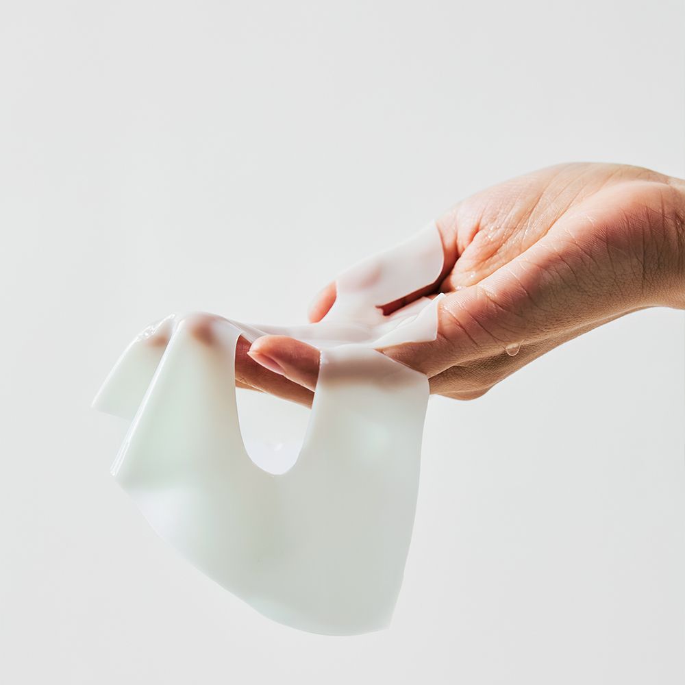 Hand hält eine weiße Gesichtsmaske. Maske ist feucht und flexibel. Keine Verpackung sichtbar.