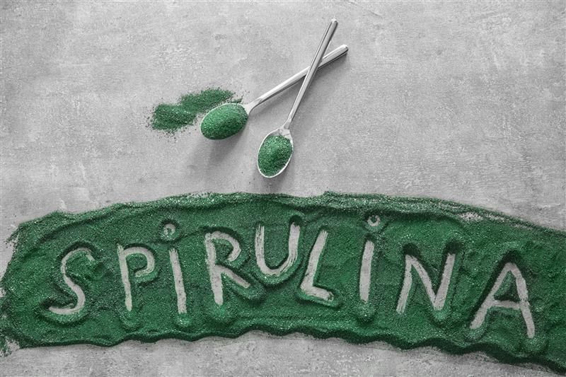 Grünes Spirulina-Pulver auf grauem Untergrund. Schriftzug "SPIRULINA" aus Pulver. Zwei Löffel mit Pulver.