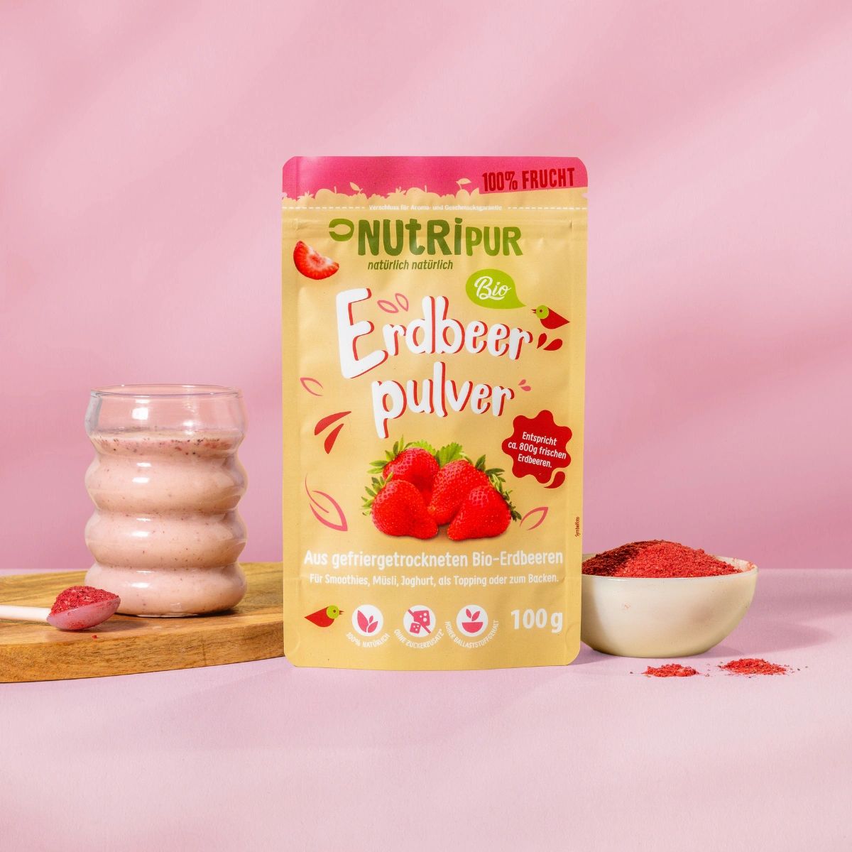 Produktpräsentation: Erdbeerpulver, Glas mit Erdbeershake, Schale mit Pulver. Auf Holzbrett. Hintergrund rosa.