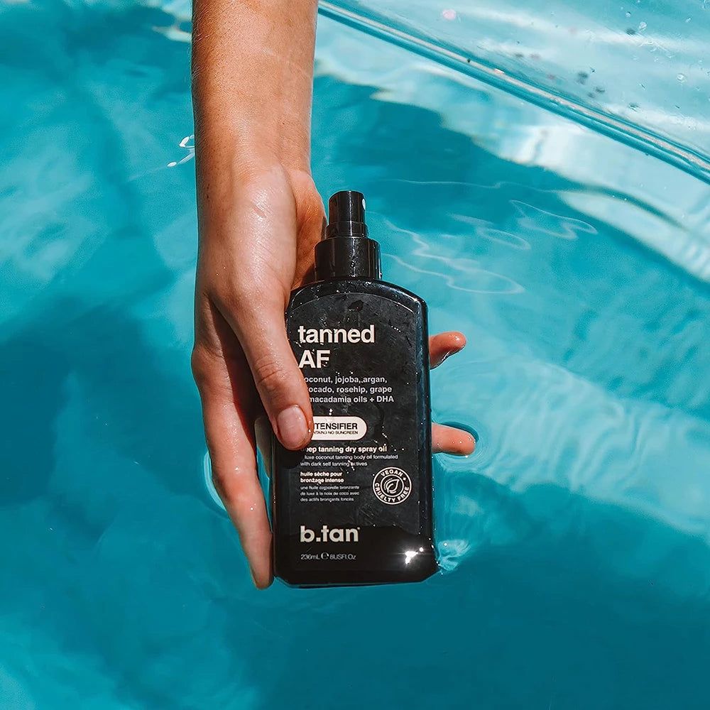 Schwarze Sprühflasche 'tanned AF' in einer Hand über blauem Wasser. 'b.tan' ist sichtbar. Enthält Kokosnuss, Jojoba, Argan, Avocado, Hagebutte, Traube & Macadamia-Öle + DHA.