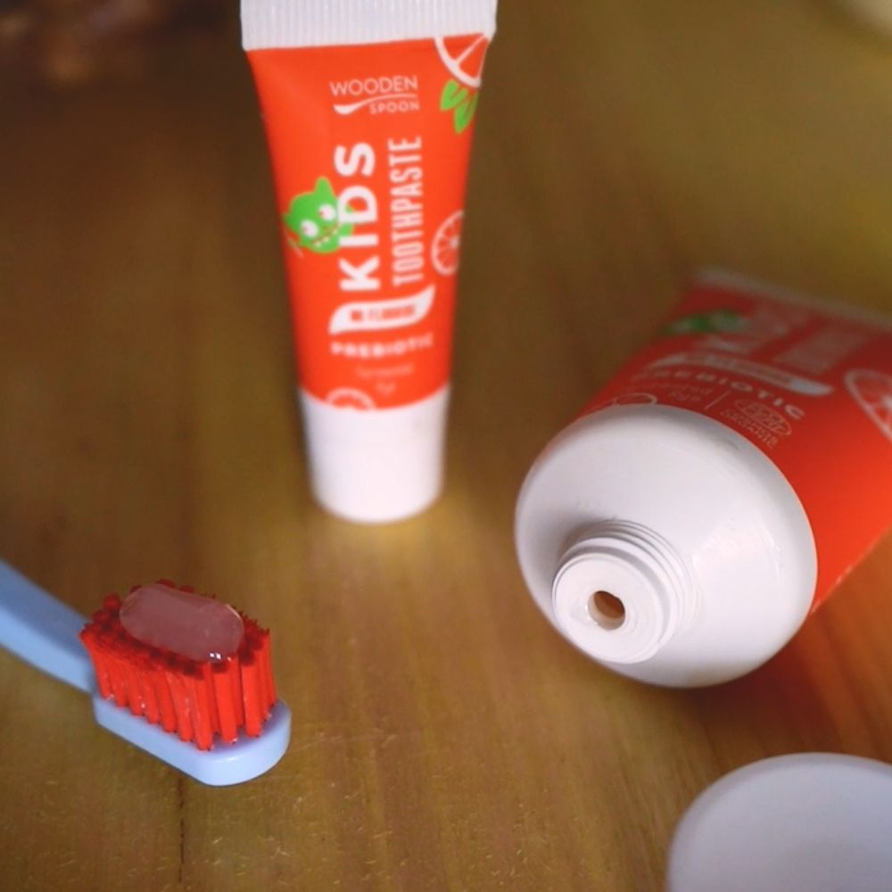Zahnbürste und zwei Tuben. Aufschrift: KIDS TOOTHPASTE, PREBIOTIC, NO FLUORIDE. Mit Illustrationen. Eine Tube geöffnet.