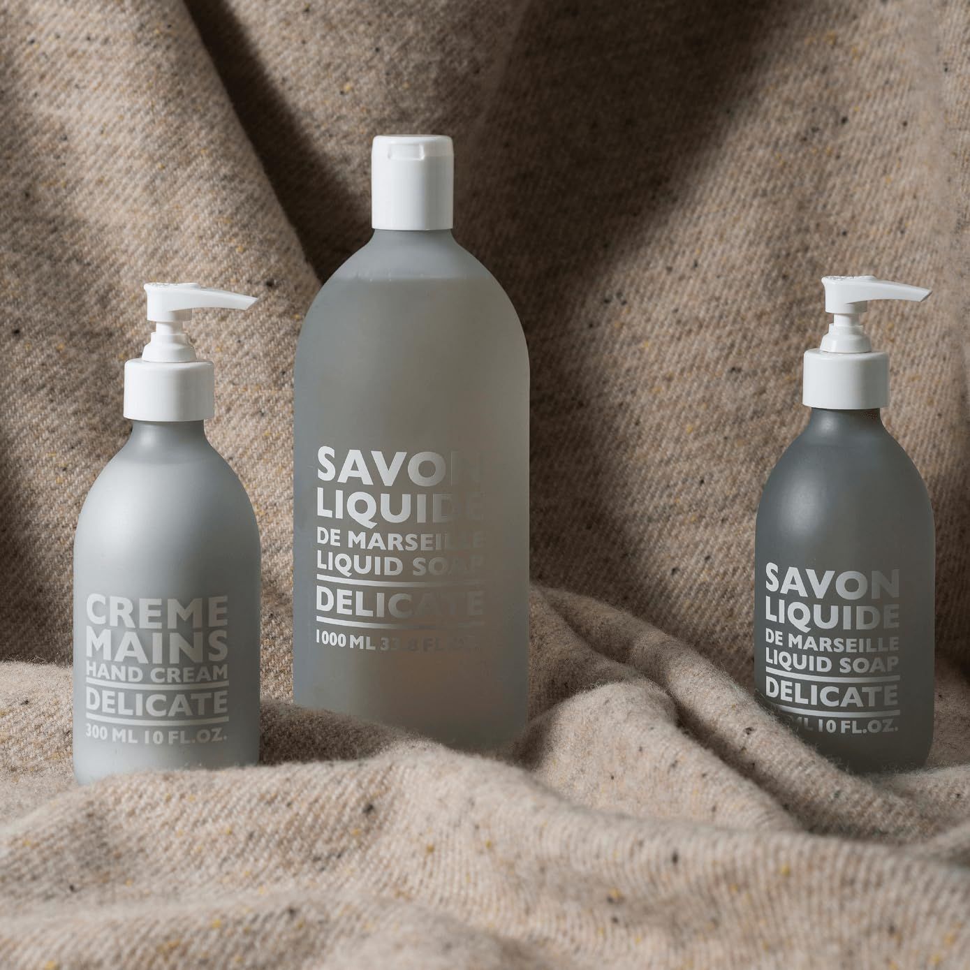 Drei Flaschen auf einem beigen Stoff. Zwei mit Pumpaufsatz, eine ohne. Text: Savon Liquide Delicate, Crème Mains Delicate.