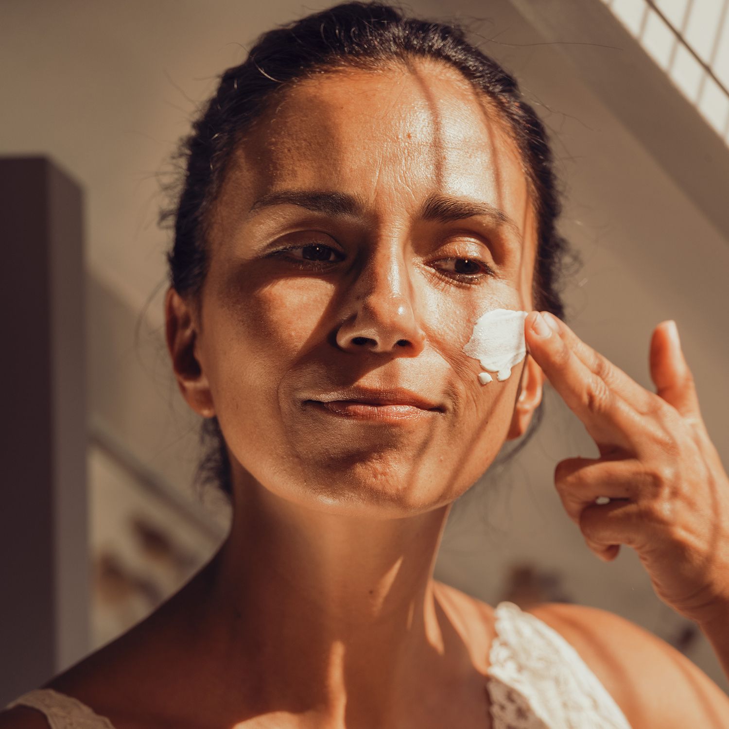 Frau trägt Sonnenfluid auf ihr Gesicht auf. Weiße Creme auf der Haut. Person trägt ein weißes Oberteil.