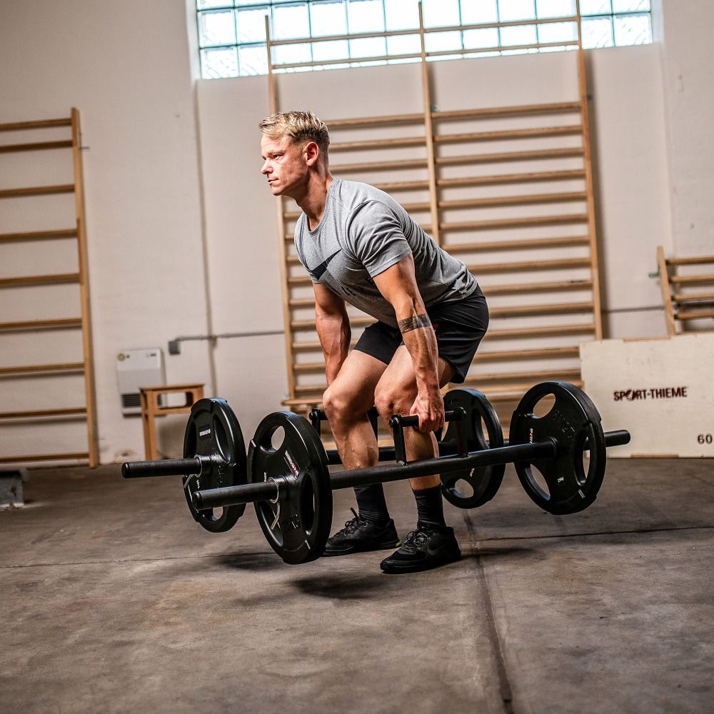 Mann hebt Farmers Walk Bar mit Gewichten. Schwarze Stange mit Griffen und Gewichtsscheiben. Sport-Thieme Logo auf Kiste.