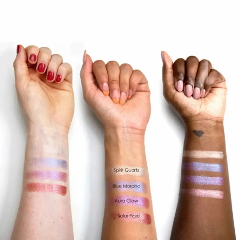 Drei Arme mit verschiedenen Hauttönen zeigen Lidschatten-Swatches. Beschriftungen: Spirit Quartz, Blue Morpho, Aura Glow, Solar Flare. Die Haut ist weiß, hellbraun und dunkelbraun.