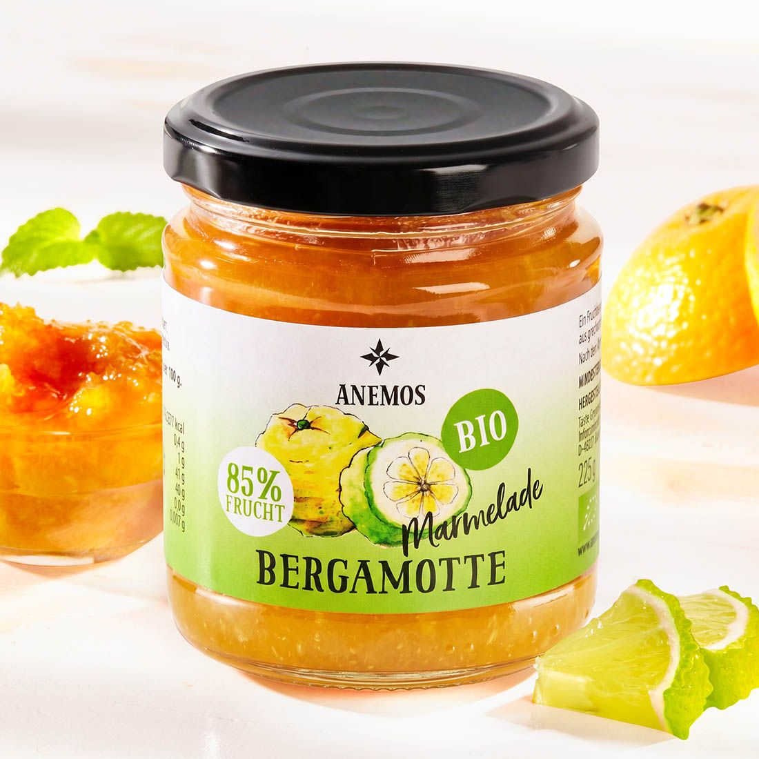 Glas mit Bergamotte-Marmelade. Etikett mit Logo, Schriftzug und Bio-Siegel. Neben dem Glas: Bergamotte-Stücke und Minzblätter.
