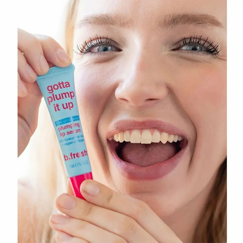 Frau hält eine blau-rosa Tube "gotta plump it up" vor ihrem Mund. Marke: b.fresh. Lippen sind leicht gerötet.