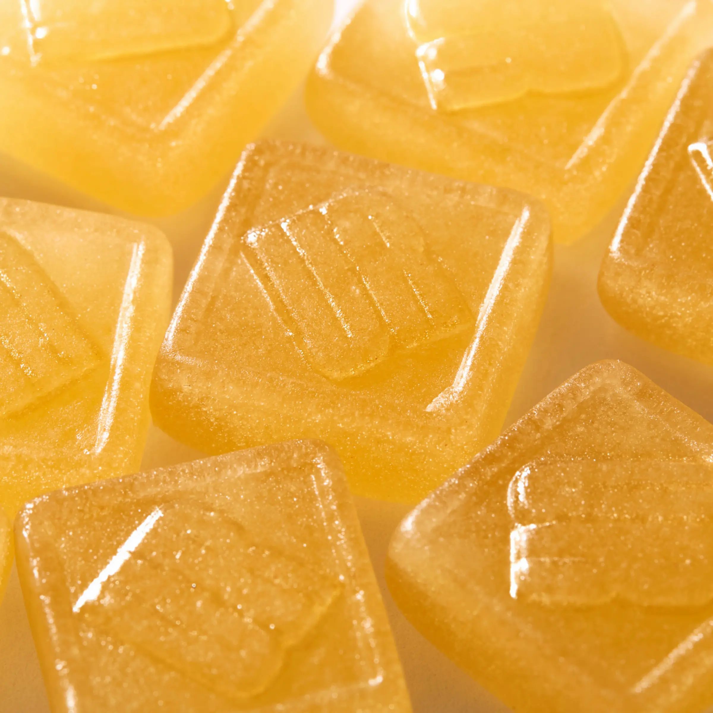 Mehrere quadratische, gelbe Gummibärchen mit einem geprägten Logo. Einige sind leicht transparent.