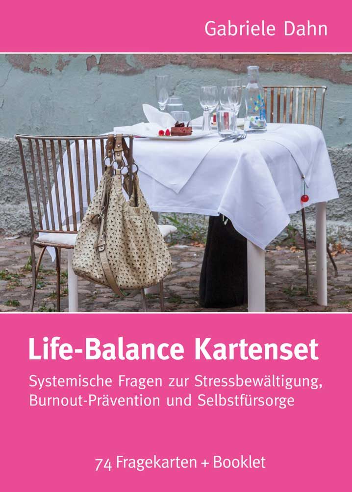 Kartenset "Life-Balance" mit 74 Fragekarten und Booklet. Rosa Hintergrund mit Text. Tisch mit Gläsern und Essen im Hintergrund.