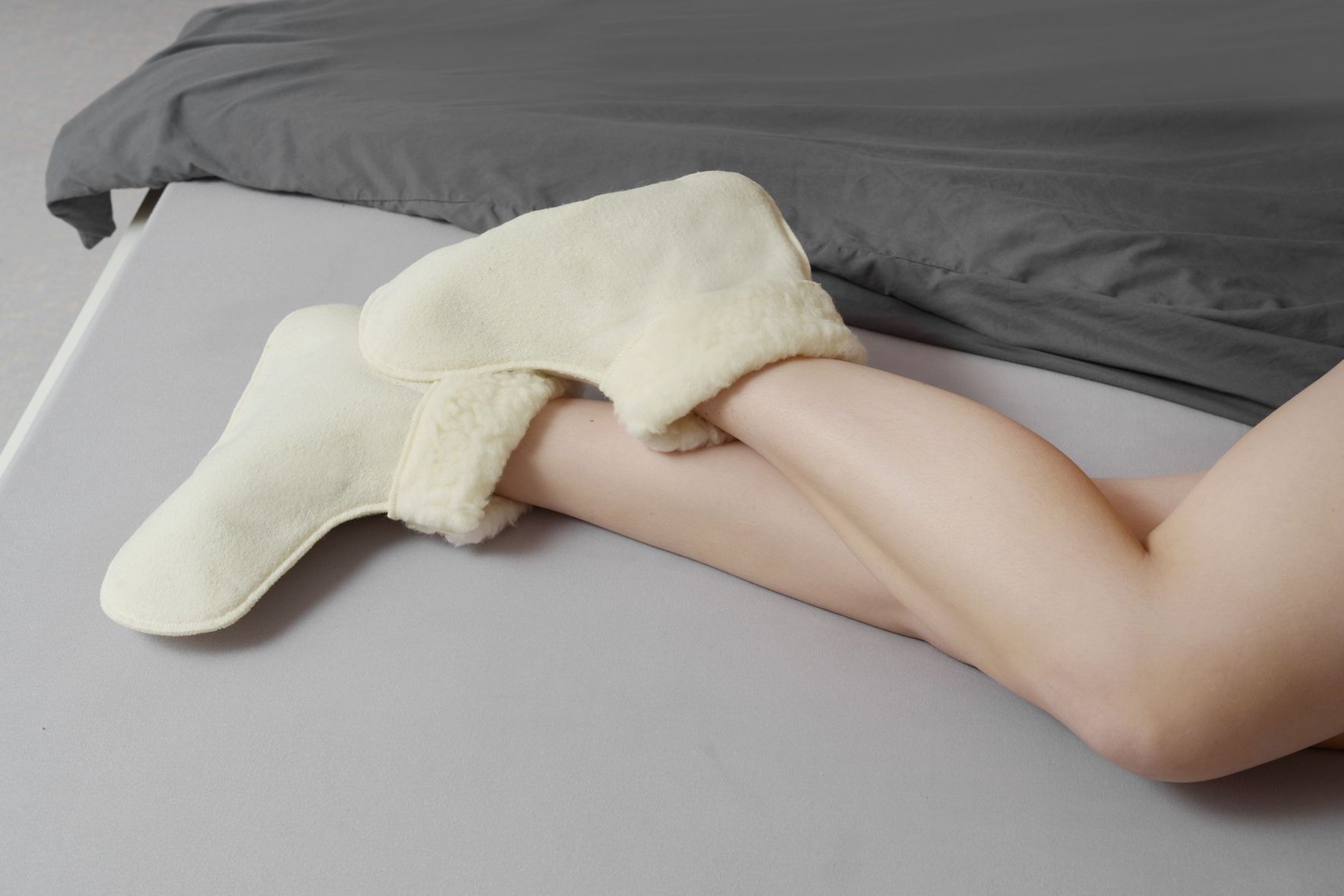 Person trägt cremefarbene Schurwoll-Bettschuhe. Füße und Unterschenkel auf Bett mit grauer Decke.