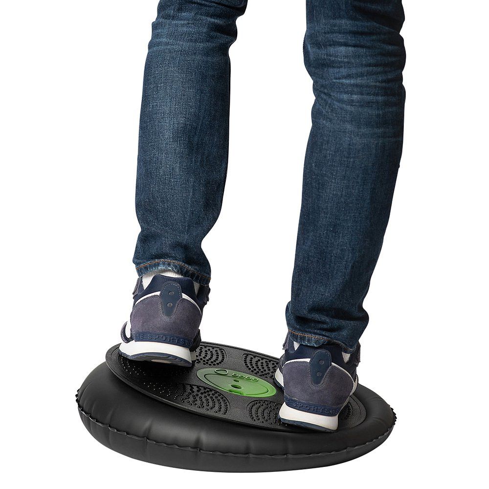 Person steht auf einem Balance Board. Schwarzes Board, grüne Mitte. Füße in Jeans und Turnschuhen.