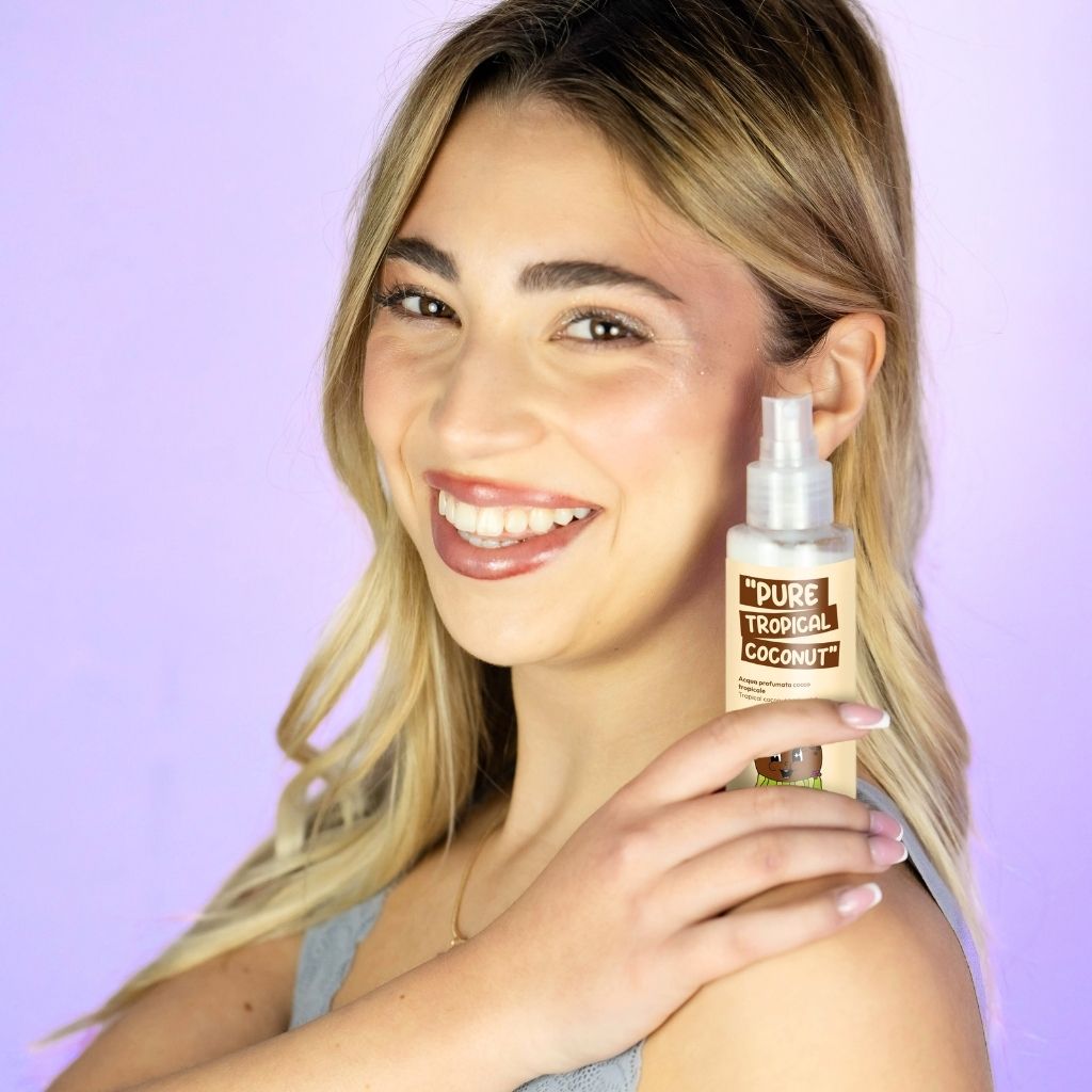 Una donna tiene in mano una bottiglia di spray per il corpo "Pure Tropical Coconut". La bottiglia è trasparente con un'etichetta marrone e un erogatore a spruzzo. La donna sorride e tiene la bottiglia davanti a sé.